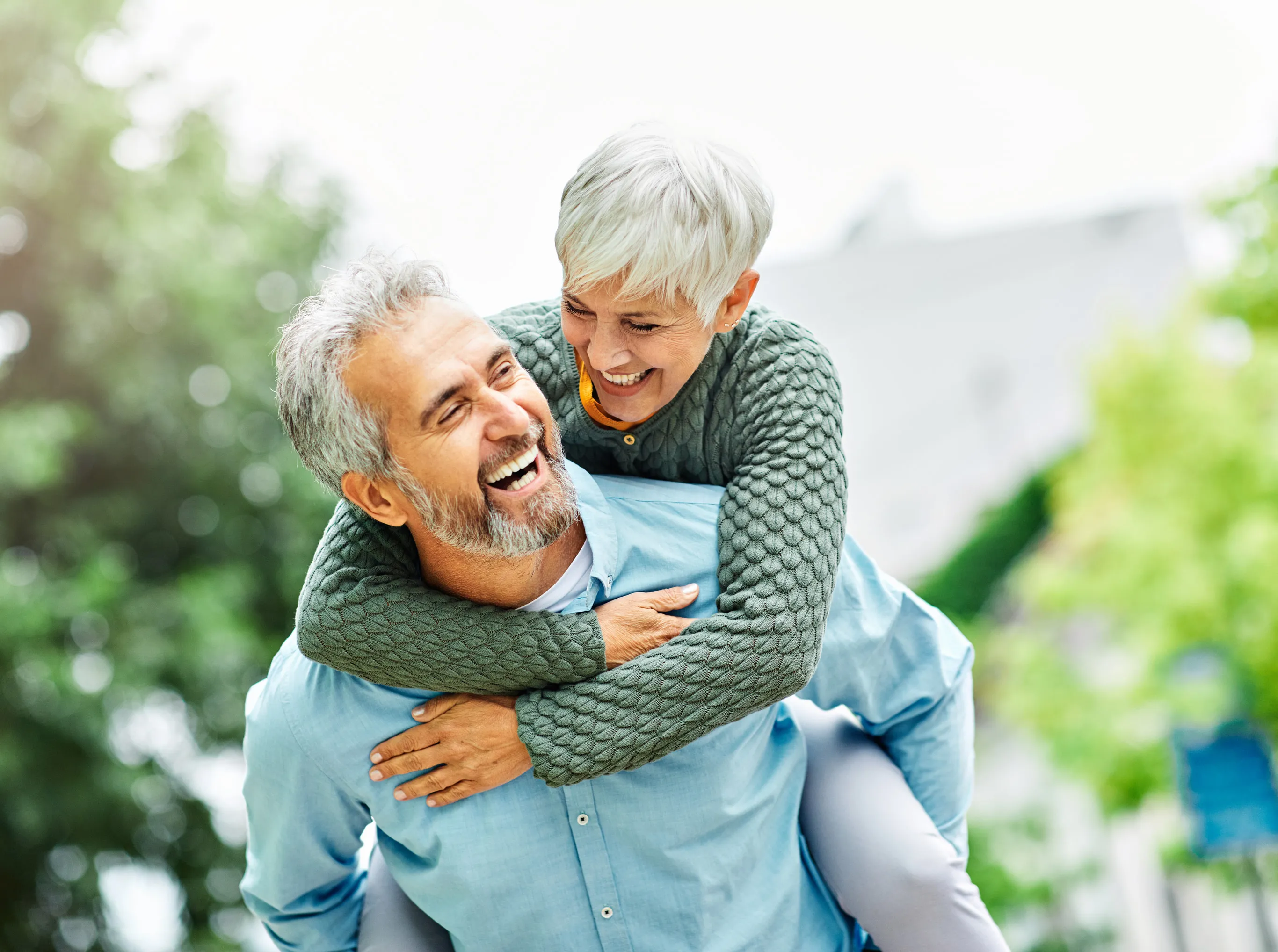 A laughing older couple outdoors, the woman riding on the man’s back, surrounded by soft green foliage.